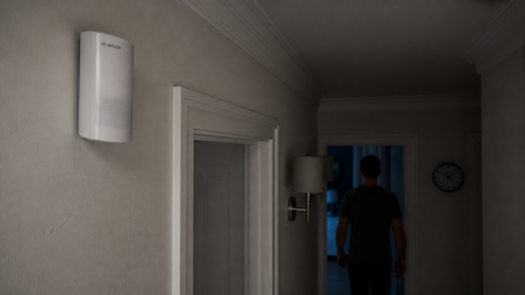 Bosch BSEN-M motion detector mounted high in a dim hallway while a person walks past and the wall sconce remains off.